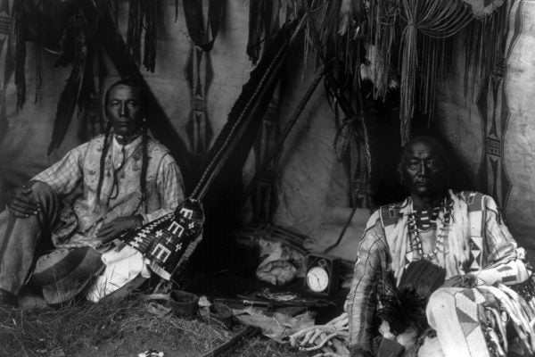 Little Plume and son Yellow Kidney seated on ground inside lodge, pipe between them in a Piegan Lodge.