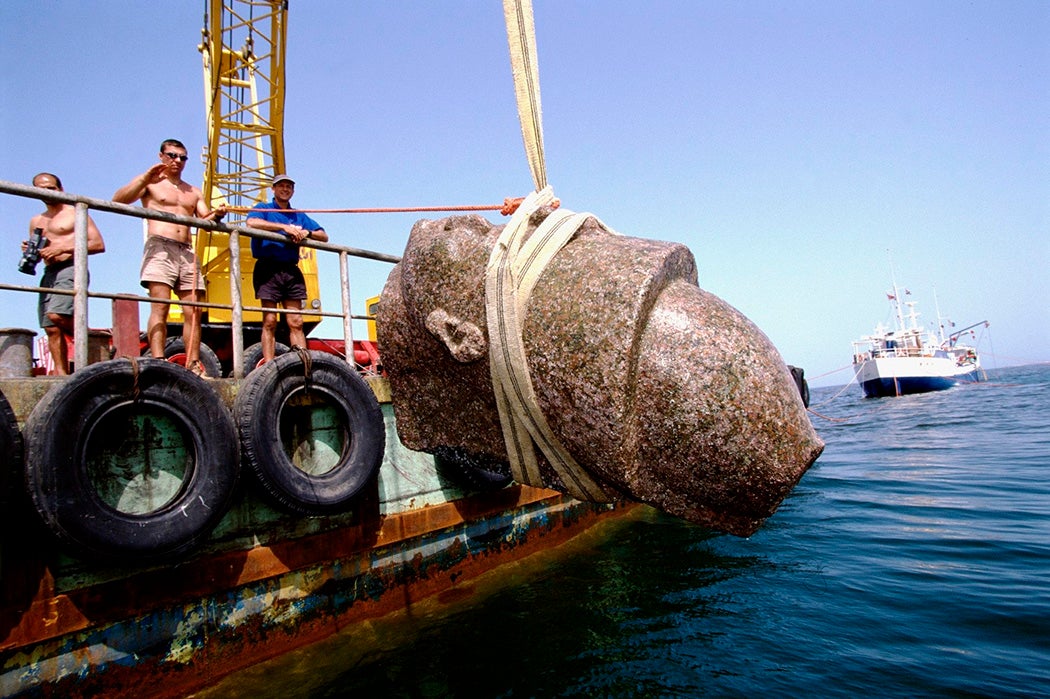 The head of a colossal red granite statue of a pharaoh is raised to the surface.