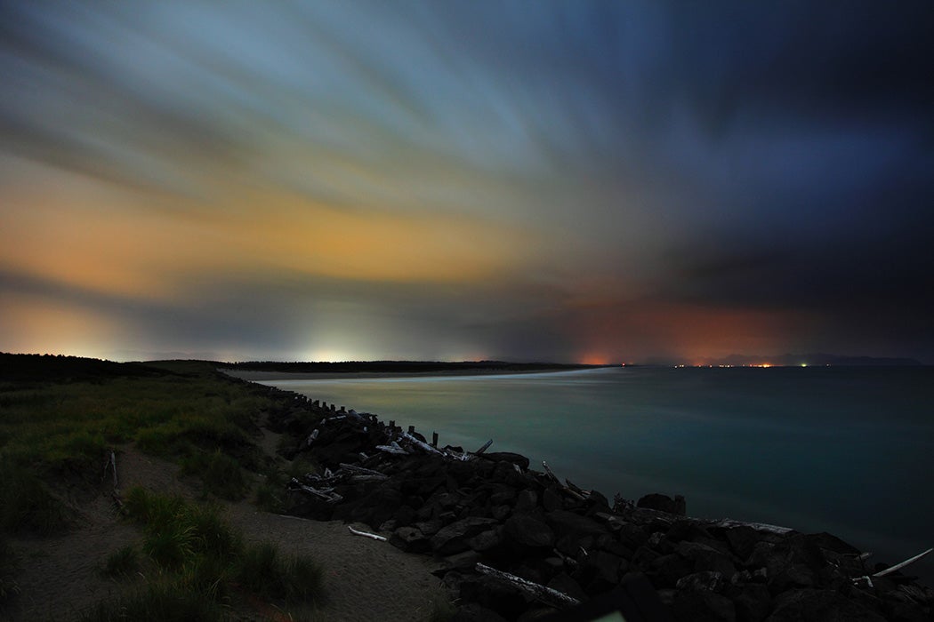 The distant lights of Astoria, Oregon can be seen lighting up the overcast, cloudy skies above the Oregon Coast near the mouth of the Columbia River