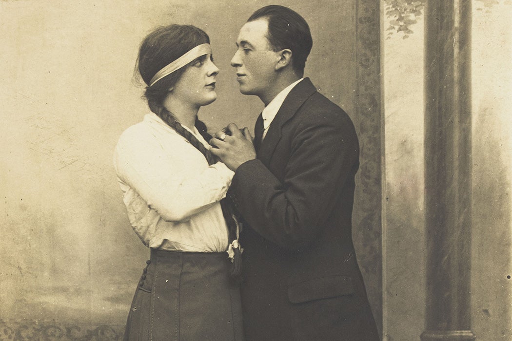 A man in drag and a man in male clothes looking into each others' eyes. Photographic postcard.